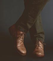A person wearing dark green pants and Scoot rugged lace-up boots by Bed Stu, handcrafted in Italy, stands with legs crossed against a dark background.