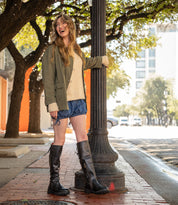 A woman stands on a city sidewalk by a lamppost, smiling and looking to the side, wearing Bed Stu Scope black lace-up back boots made from vegetable-tanned leather, a green jacket, beige sweater, and blue shorts.