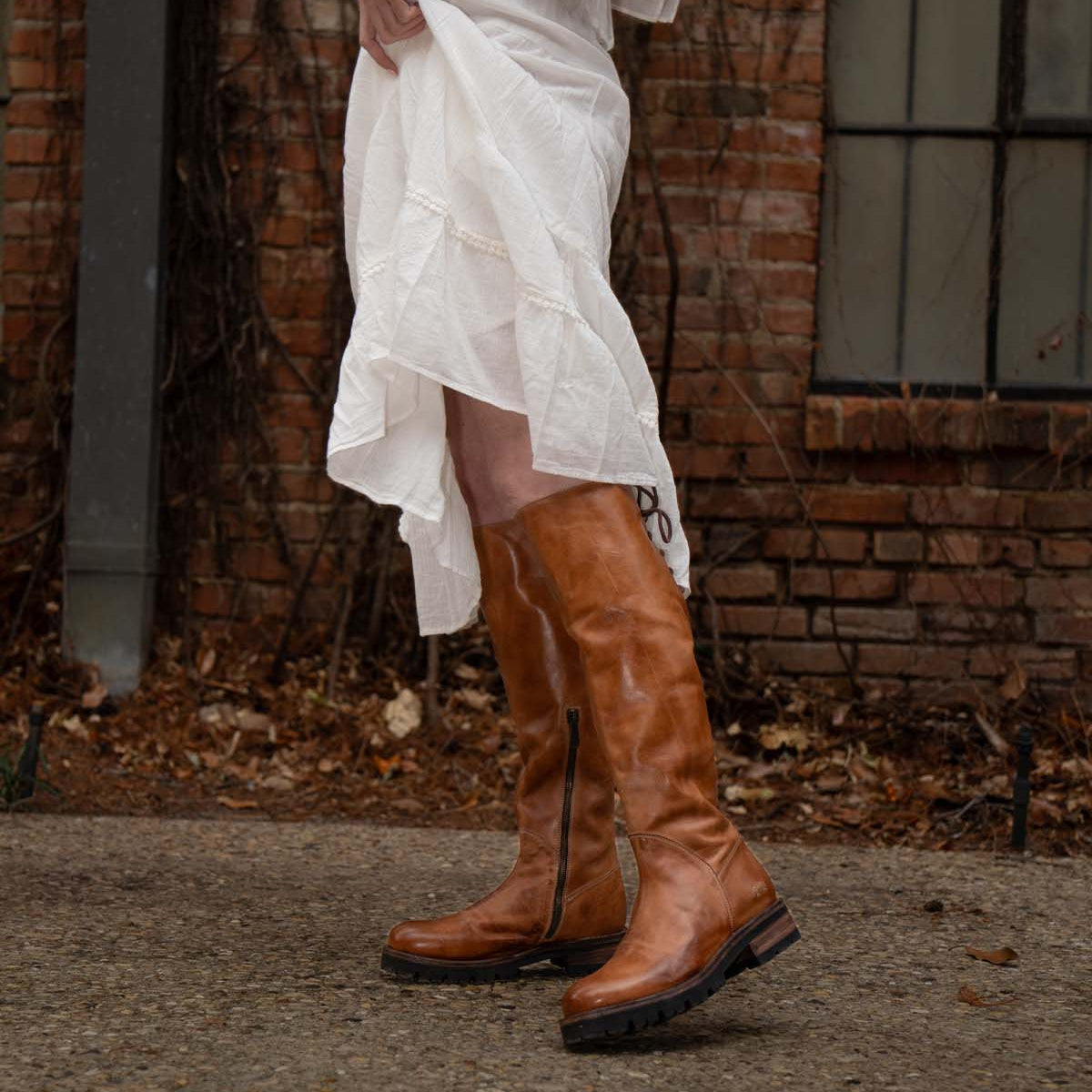 A person in a white dress lifts the hem to reveal Bed Stu Scope tall women’s leather boots with a lace-up back, standing on a stone path by a brick wall and window.