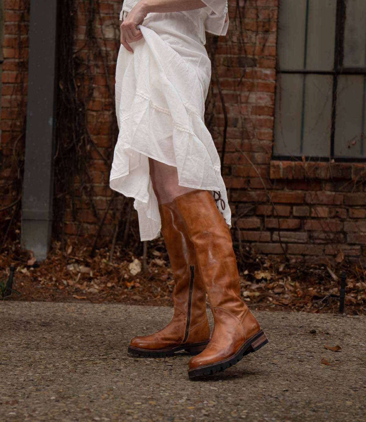 A person in a white dress lifts the hem to reveal Bed Stu Scope tall women’s leather boots with a lace-up back, standing on a stone path by a brick wall and window.