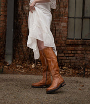 A person in a white dress lifts the hem to reveal Bed Stu Scope tall women’s leather boots with a lace-up back, standing on a stone path by a brick wall and window.