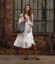 A woman in a white dress and tall leather boots stands before a brick wall with large windows, carrying the Bed Stu Scope, a large dark brown vegetable-tanned leather shoulder bag.