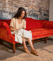 A woman in a beige top and white skirt sits on an orange velvet couch against a brick wall, gazing at her Bed Stu Scurry sustainable women's shoe made from vegetable-tanned leather, hands gently resting in her lap.