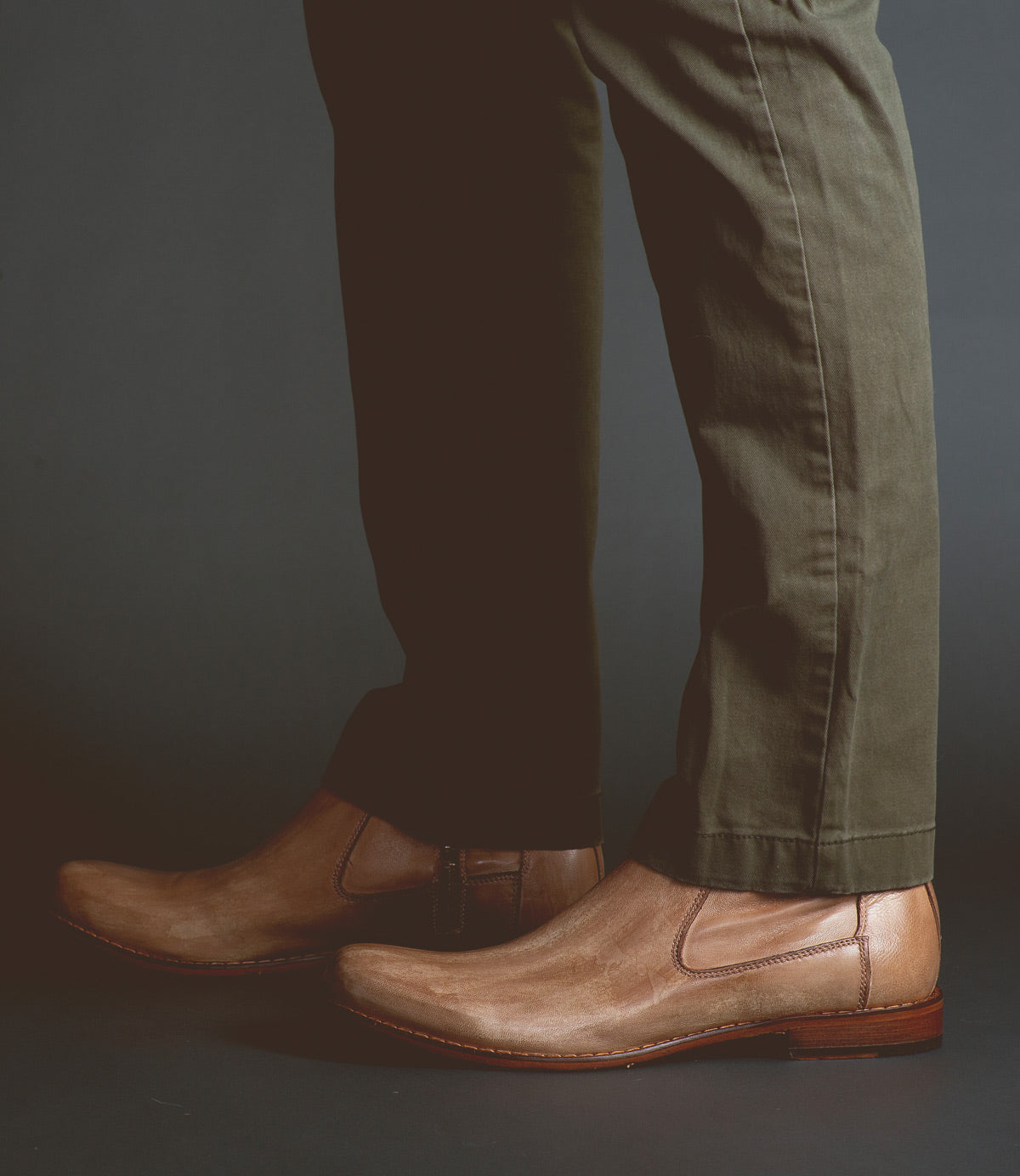 A person wearing olive green pants and brown vegetable-tanned leather slip-on shoes crafted with artisanal craftsmanship, specifically the Seek by Bed Stu, against a dark background.