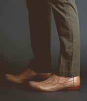 A person wearing olive green pants and brown vegetable-tanned leather slip-on shoes crafted with artisanal craftsmanship, specifically the Seek by Bed Stu, against a dark background.