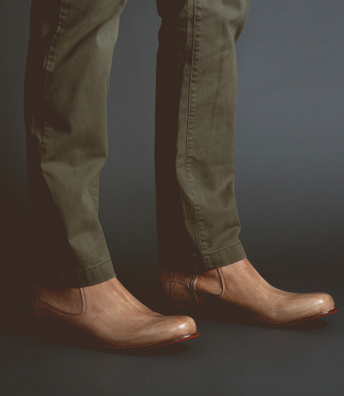 A person wearing olive green pants and light brown vegetable-tanned Bed Stu Seek slip-on shoes, showcasing artisanal craftsmanship, stands against a dark background.