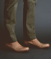 A person wearing olive green pants and light brown vegetable-tanned Bed Stu Seek slip-on shoes, showcasing artisanal craftsmanship, stands against a dark background.