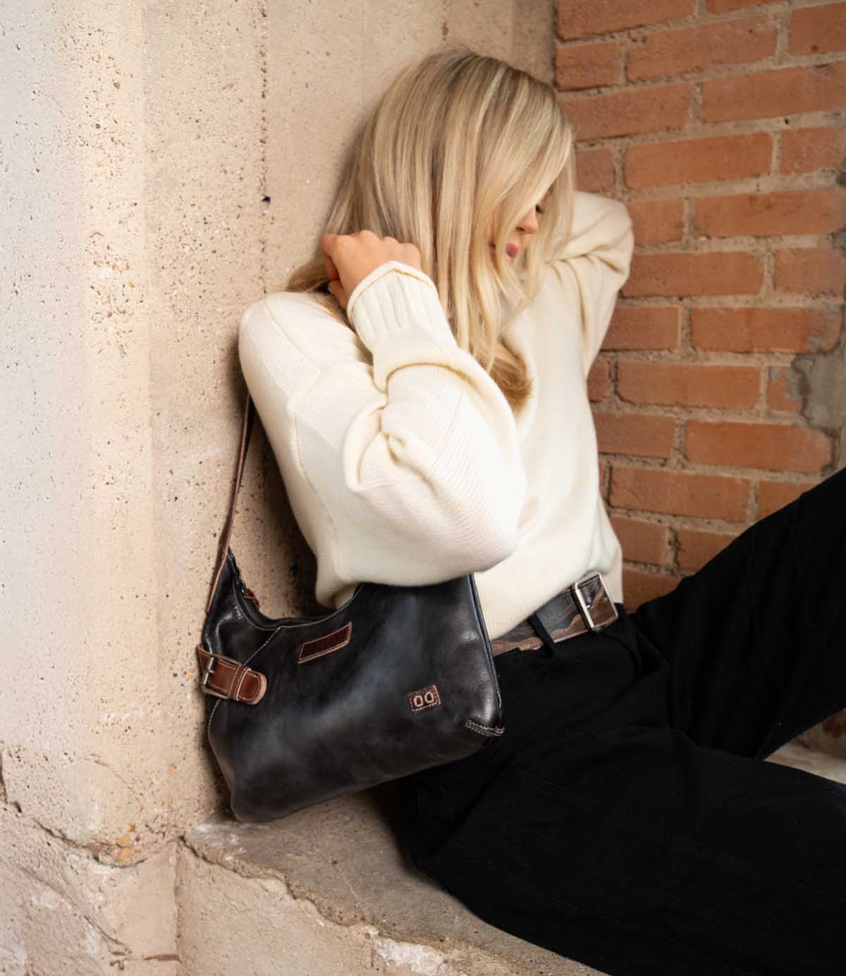 A person with blonde hair sits against a brick wall, wearing a cream sweater and black pants, holding the Bed|Stü Seemly—a compact black shoulder bag with brown accents. Stylish and versatile, Seemly is the perfect accessory for everyday looks.