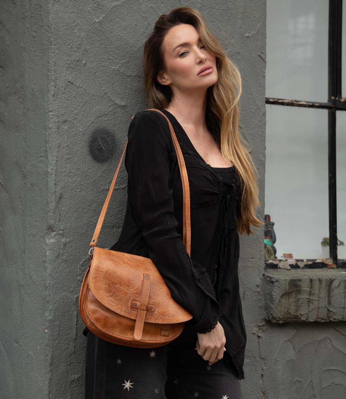 A woman with long hair stands against a textured gray wall, wearing a black outfit and carrying the Bed Stu Sequoia EM tan crossbody saddle bag with an adjustable strap.