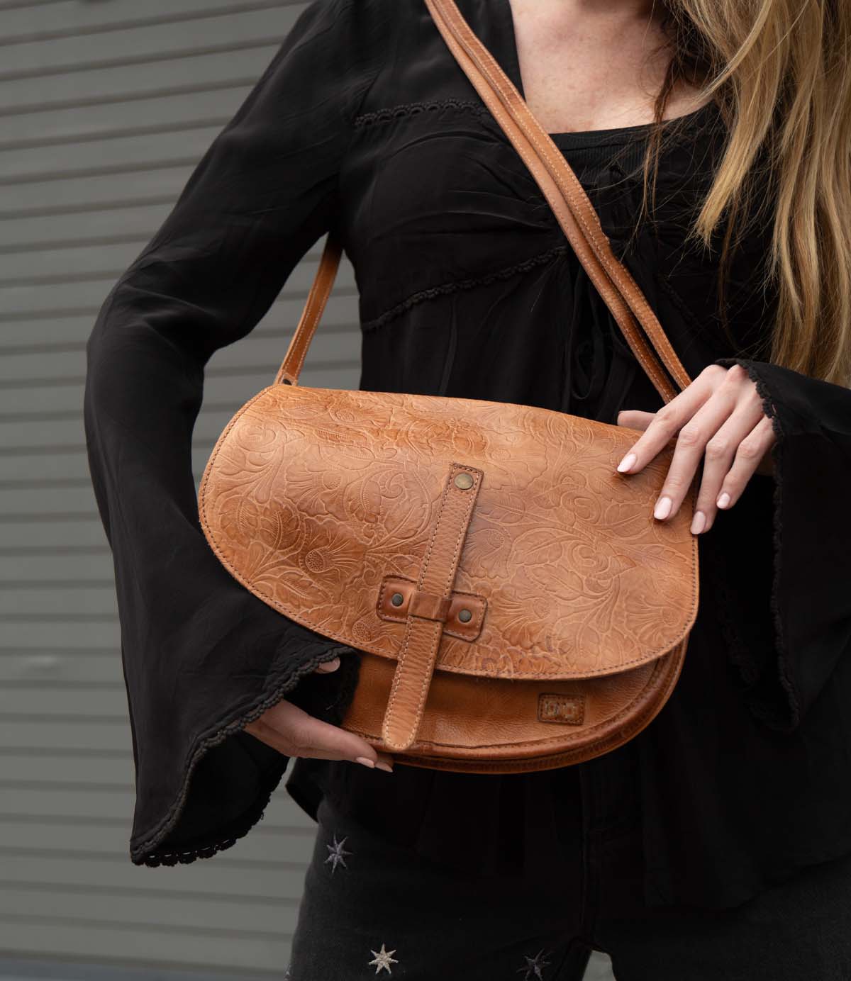 A woman in a black long-sleeve top holds the Bed Stu Sequoia EM, a tan vegetable-tanned leather shoulder bag with embossed patterns and an adjustable crossbody strap.
