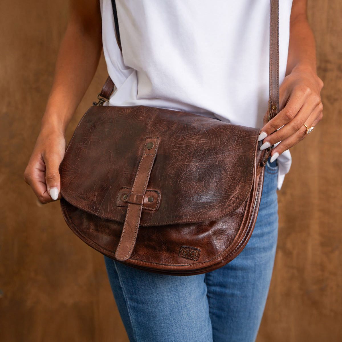 A person in a white shirt and blue jeans holds the Bed Stu Sequoia EM, a vegetable-tanned leather crossbody saddle bag with a front strap closure.