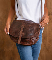 A person in a white shirt and blue jeans holds the Bed Stu Sequoia EM, a vegetable-tanned leather crossbody saddle bag with a front strap closure.