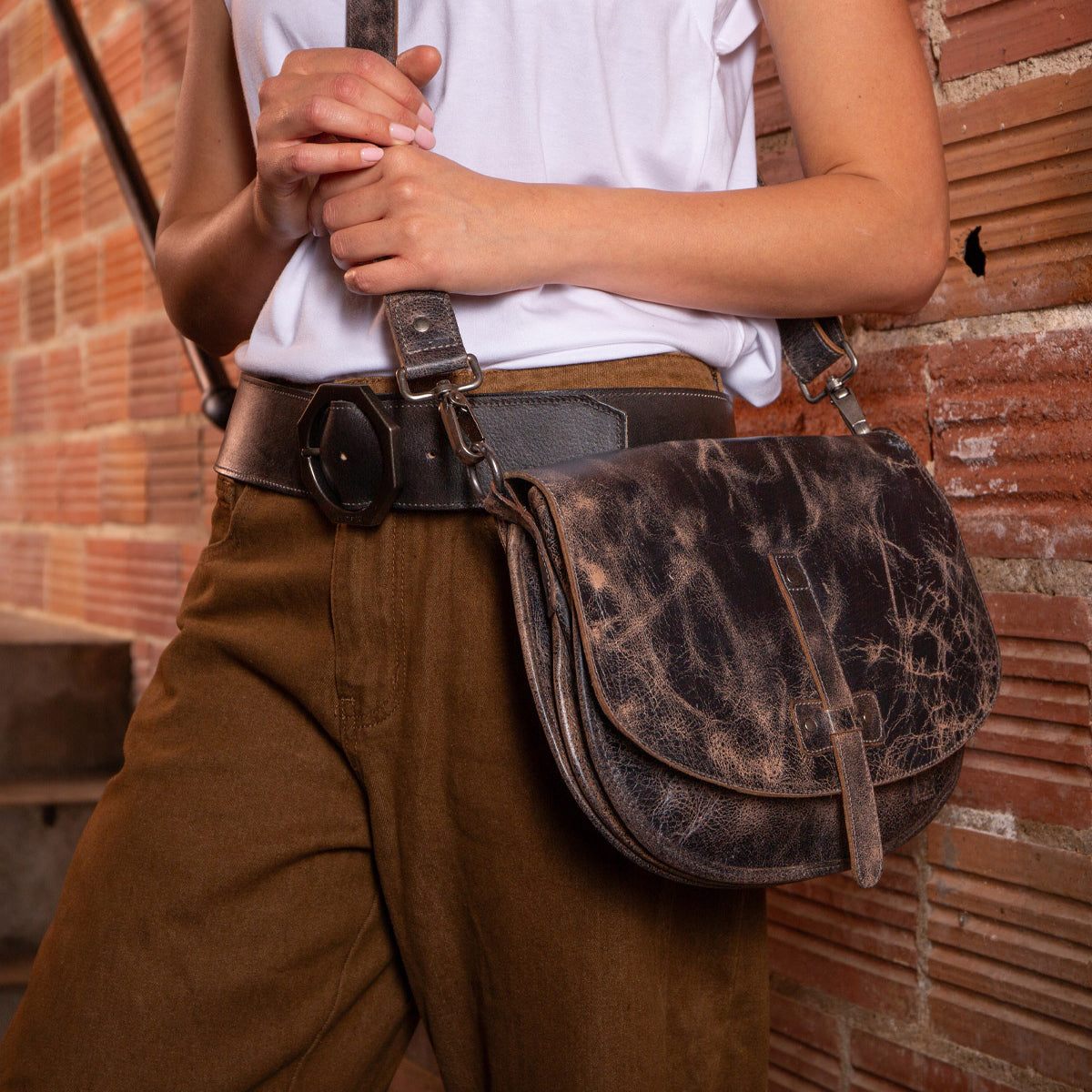 A person in brown pants and a white shirt holds the Bed Stu Sequoia, a distressed brown vegetable-tanned leather crossbody bag with an adjustable strap, standing in front of a brick wall.