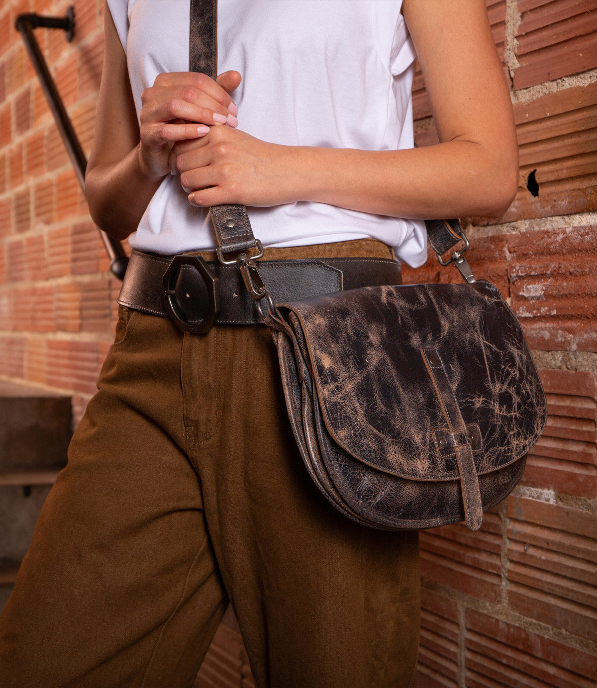 A person in brown pants and a white shirt holds the Bed Stu Sequoia, a distressed brown vegetable-tanned leather crossbody bag with an adjustable strap, standing in front of a brick wall.