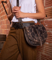 A person in brown pants and a white shirt holds the Bed Stu Sequoia, a distressed brown vegetable-tanned leather crossbody bag with an adjustable strap, standing in front of a brick wall.