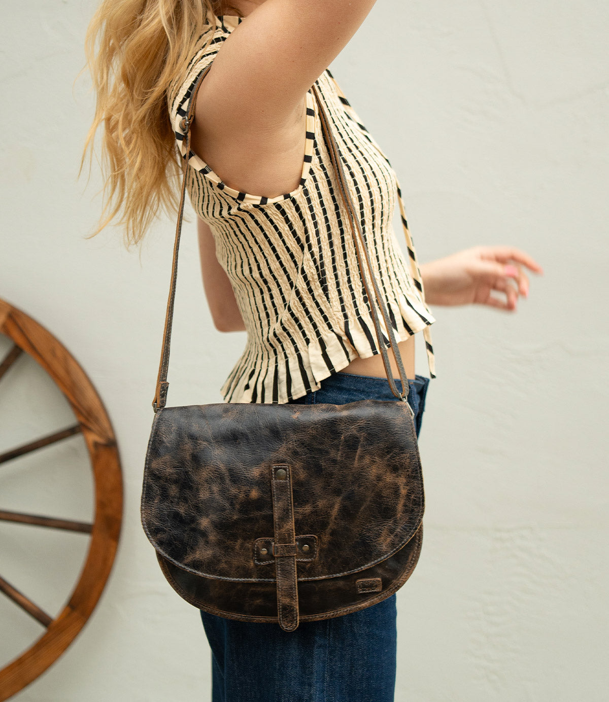 A person wearing a sleeveless striped top and jeans holds their arm up, carrying the Bed|Stü Sequoia—a worn brown vegetable-tanned leather crossbody bag with a sturdy strap—while a wooden wheel sits in the background.