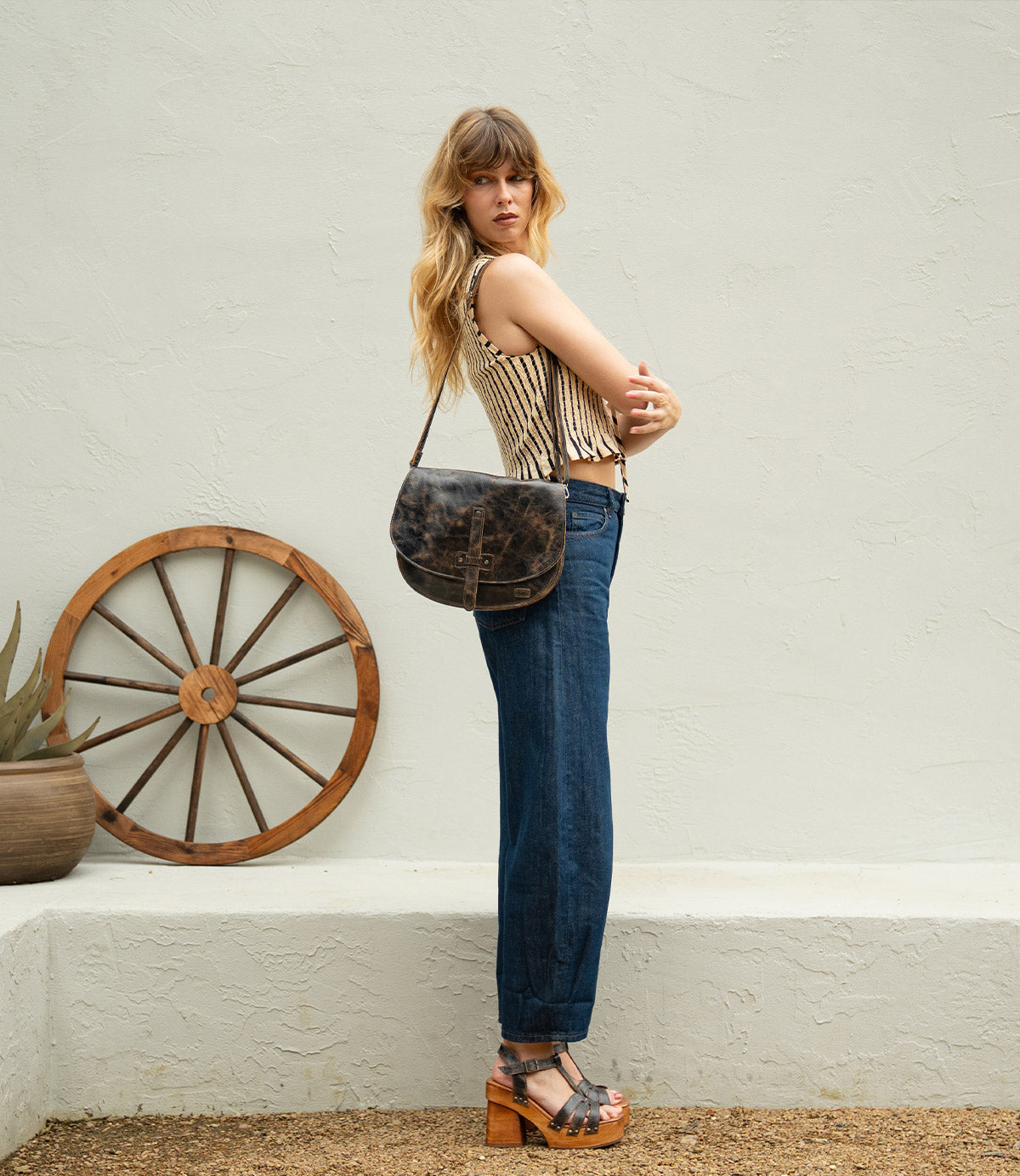 A woman stands sideways outdoors, wearing a striped sleeveless top, high-waisted jeans, platform sandals, and carrying the Bed|Stü Sequoia crossbody bag, featuring a vegetable-tanned leather finish and adjustable strap. A wooden wagon wheel and potted plant appear in the background.