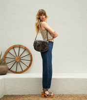 A woman stands sideways outdoors, wearing a striped sleeveless top, high-waisted jeans, platform sandals, and carrying the Bed|Stü Sequoia crossbody bag, featuring a vegetable-tanned leather finish and adjustable strap. A wooden wagon wheel and potted plant appear in the background.