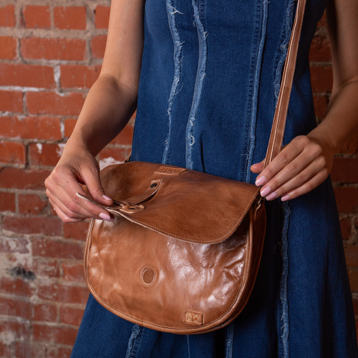 A person in a blue denim dress stands against a brick wall, holding the Bed Stu Sequoia brown vegetable-tanned leather crossbody bag with an adjustable strap and flap closure.