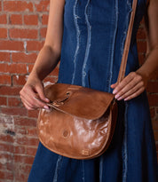 A person in a blue denim dress stands against a brick wall, holding the Bed Stu Sequoia brown vegetable-tanned leather crossbody bag with an adjustable strap and flap closure.
