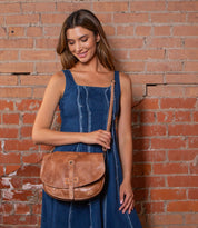 A woman in a sleeveless blue dress stands against a brick wall, holding the Bed Stu Sequoia— a brown vegetable-tanned leather crossbody bag with adjustable strap and flap closure.