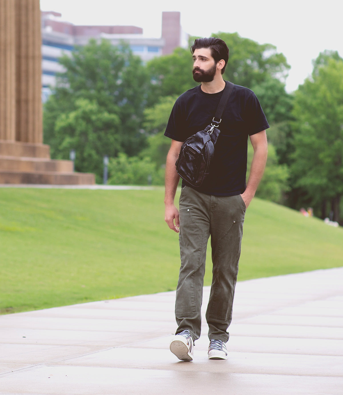 A man with a beard walks on a sidewalk in an outdoor urban park. He is wearing a black t-shirt, green pants, and white sneakers, and he carries a high-quality black crossbody bag from the Bed|Stü Men's Shop the Look' Bundle Package Price!