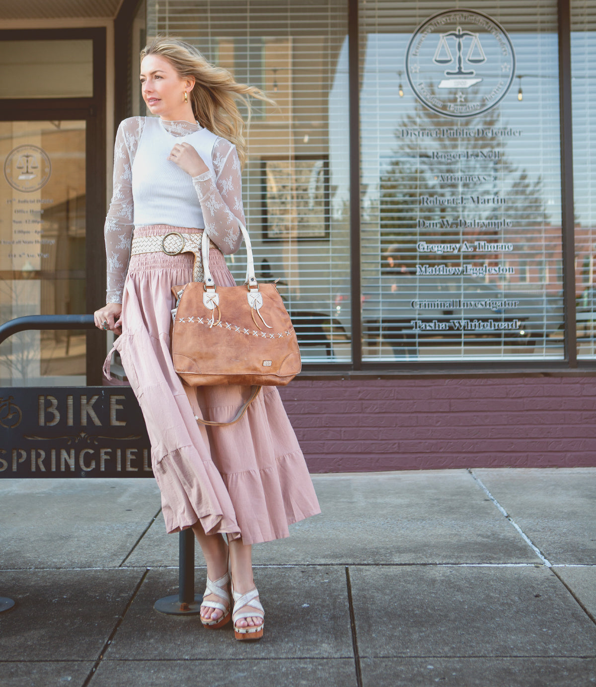 A woman in a flowing pink dress and white lace cardigan stands outside a shop, holding a large brown leather bag from Bed|Stü's 'Complete Your Look with Our 'Shop the Look' Bundle!'.