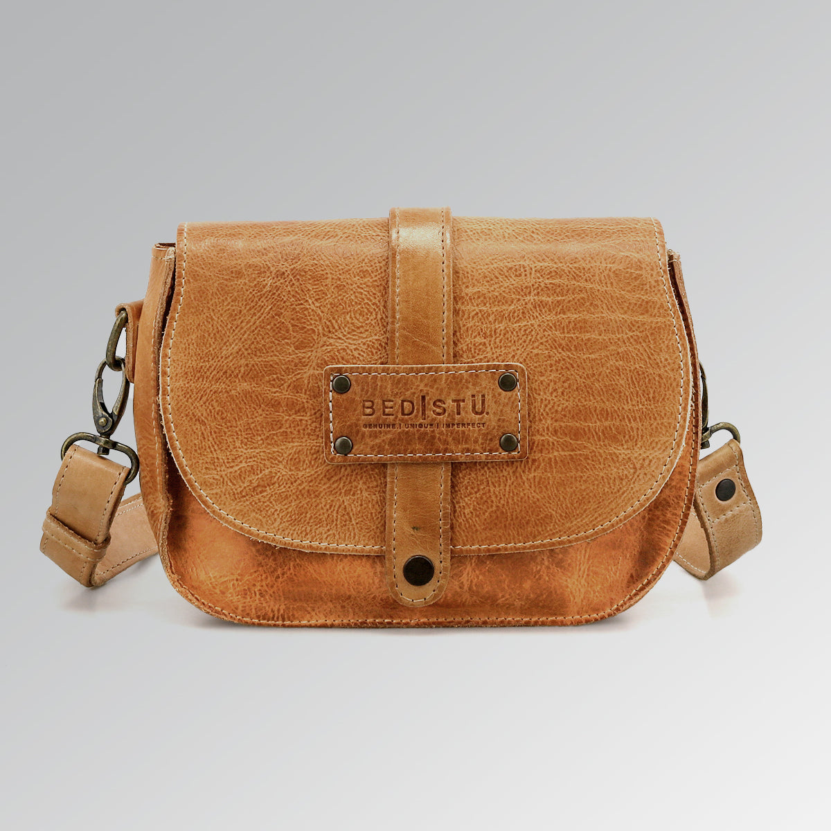 The Studio Shoreline II Body by Bed Stu is a small tan leather crossbody bag with a front flap and metal rivets, standing out with its unique style against a plain gray background.