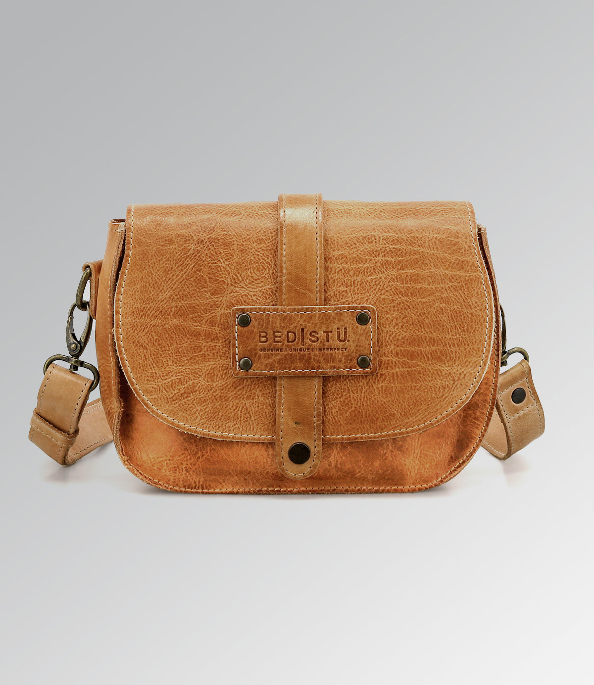 The Studio Shoreline II Body by Bed Stu is a small tan leather crossbody bag with a front flap and metal rivets, standing out with its unique style against a plain gray background.