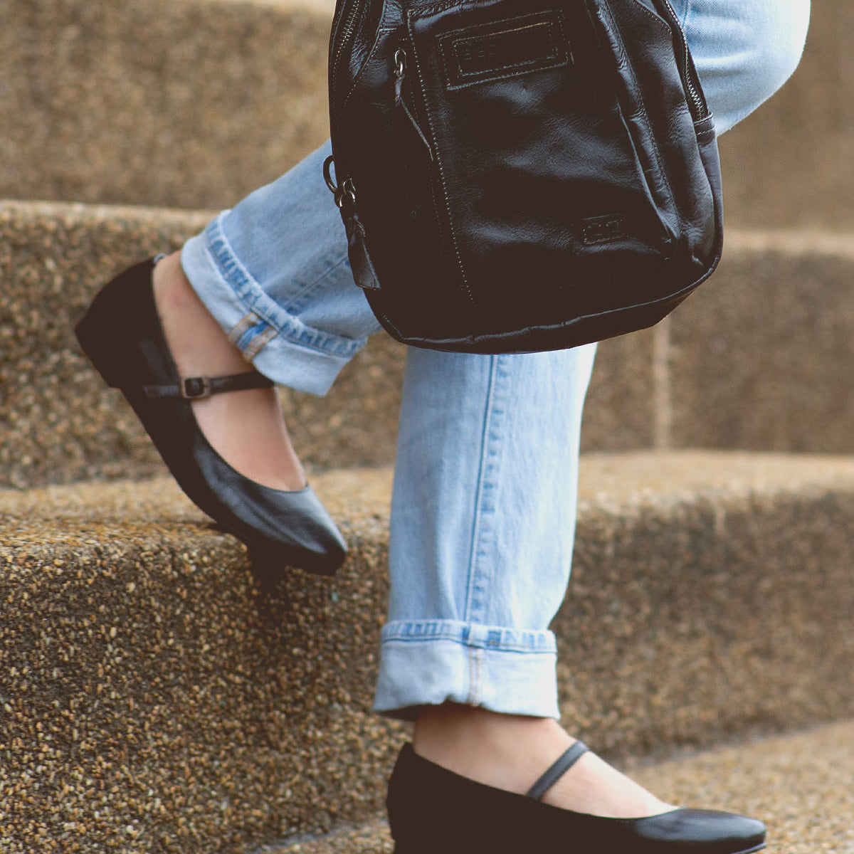 Person wearing black ballet flats and blue jeans carrying a Shuffle III by Bed Stu while walking up concrete steps, effortlessly embodying the balletic elegance of the ballerinacore trend.