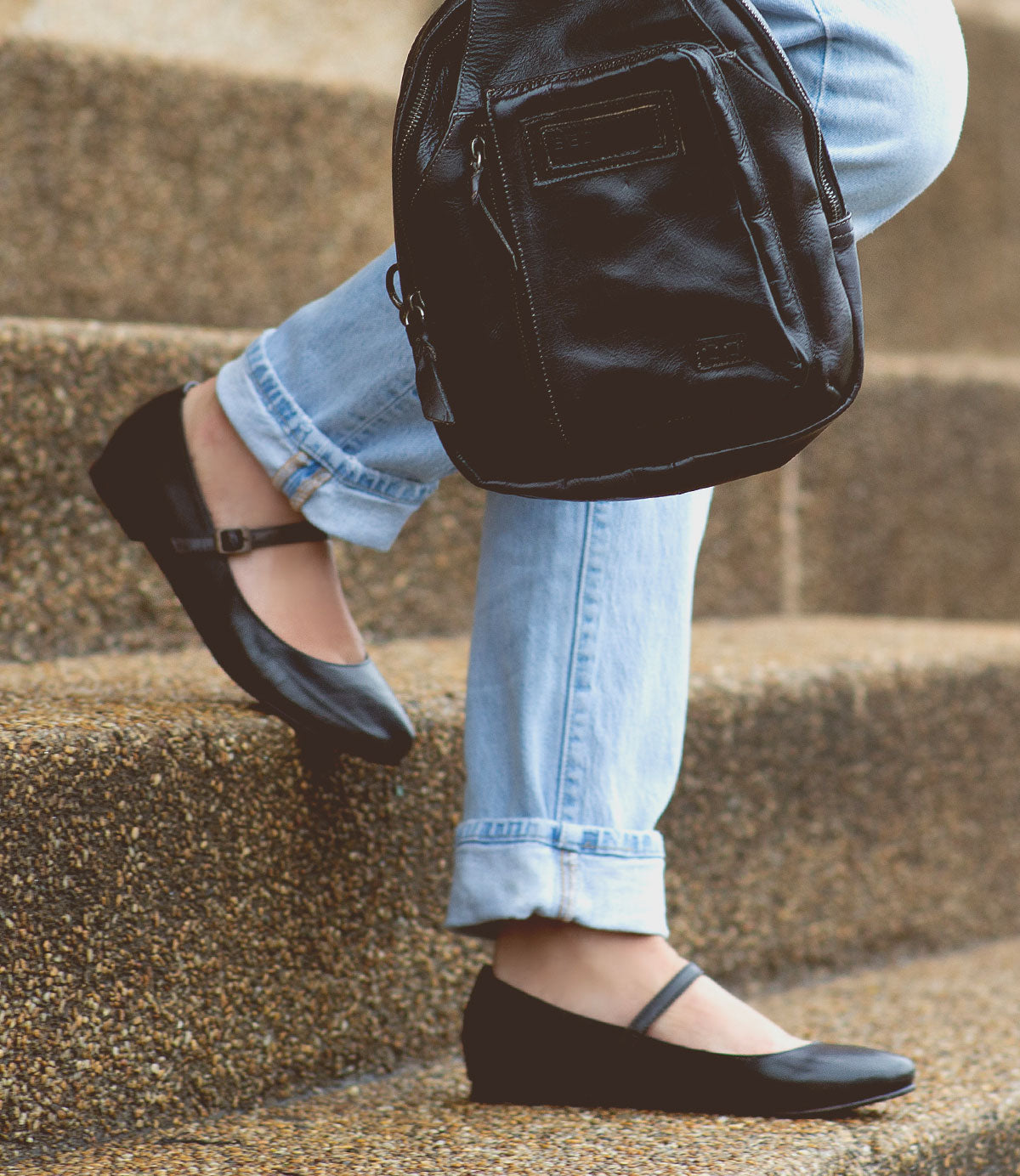 Person wearing black ballet flats and blue jeans carrying a Shuffle III by Bed Stu while walking up concrete steps, effortlessly embodying the balletic elegance of the ballerinacore trend.