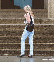 A person with long hair wearing a white top, light blue jeans, and black ballet flats is walking outside on steps, carrying a black Bed Stu Shuffle III bag.