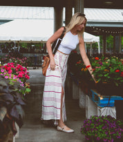 A woman in a white tank top with adjustable straps and a long striped skirt, embodying the ballerinacore trend, is reaching for flowers at a market in her Bed Stu Shuffle III shoes.