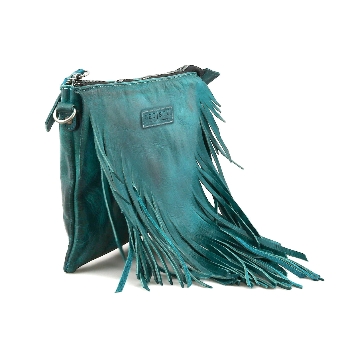The Bed Stu Silverrock is a teal leather crossbody clutch featuring a zipper closure, decorative fringe, and a small rectangular patch on the front.