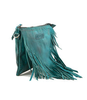 The Bed Stu Silverrock is a teal leather crossbody clutch featuring a zipper closure, decorative fringe, and a small rectangular patch on the front.