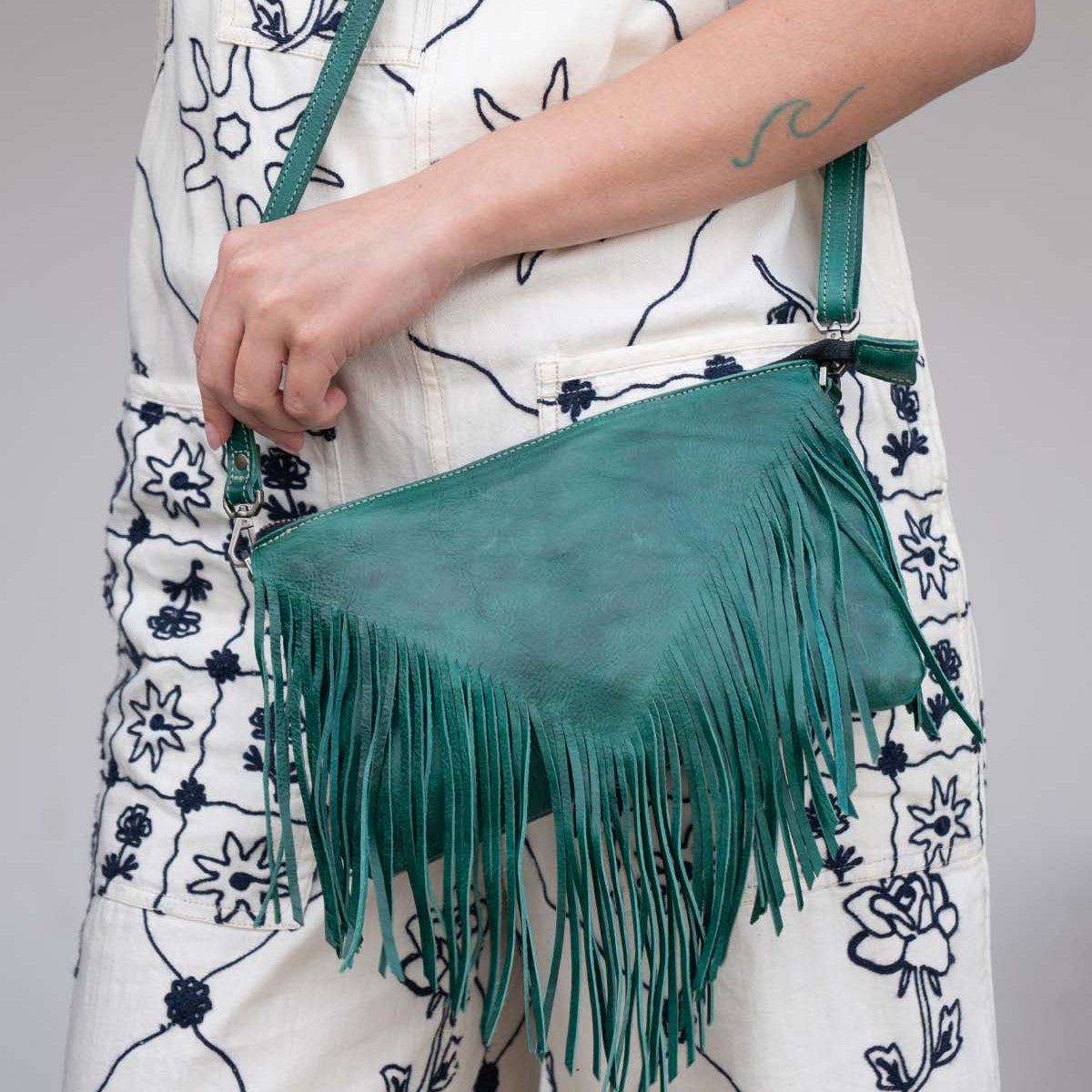 A person holds the Bed Stu Silverrock green leather fringe flap bag with crossbody strap, styled with a white jumpsuit featuring black floral patterns.