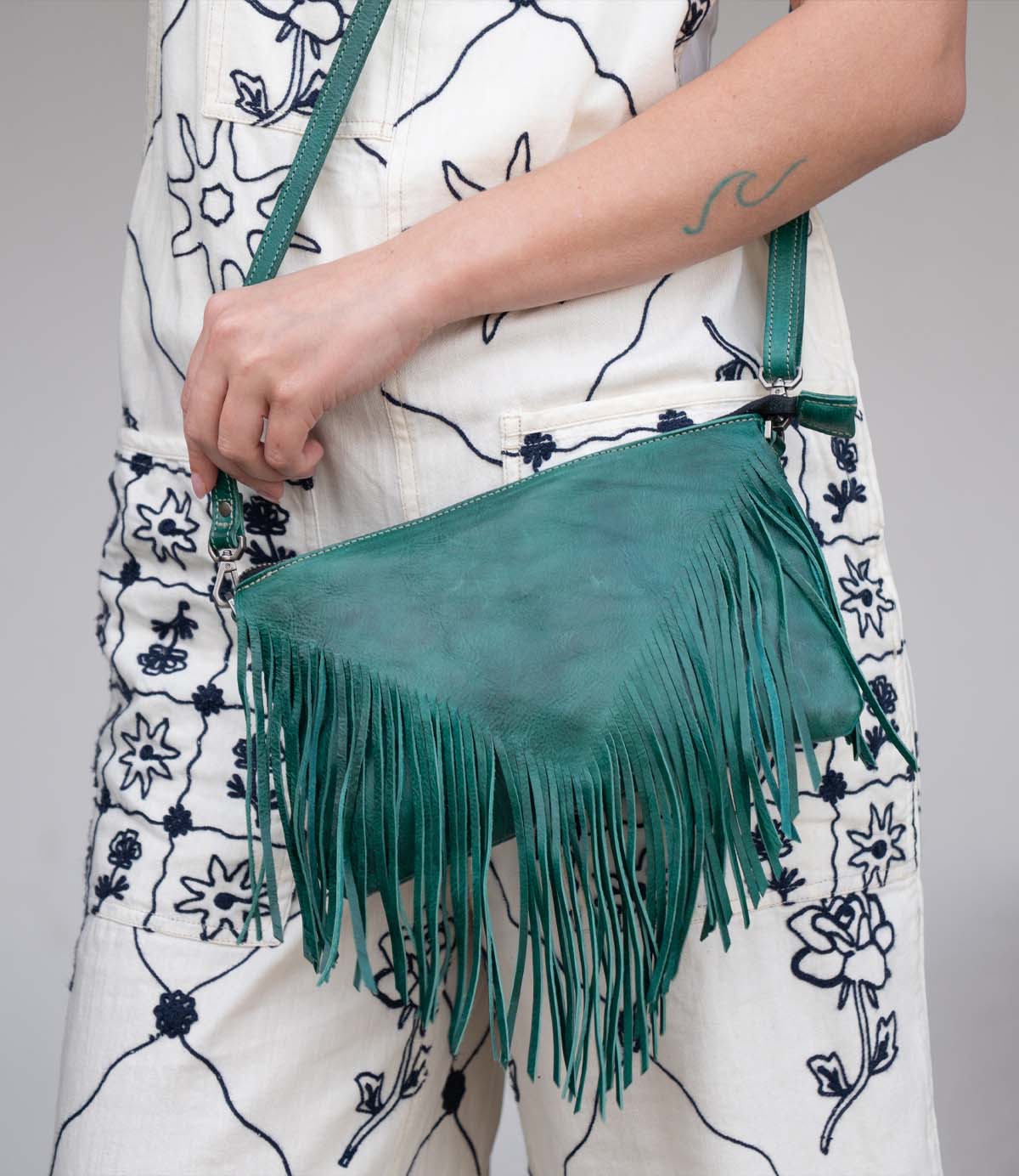 A person holds the Bed Stu Silverrock green leather fringe flap bag with crossbody strap, styled with a white jumpsuit featuring black floral patterns.