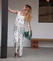 A woman in patterned white overalls and sandals stands by a wooden post, holding the Bed Stu Silverrock green crossbody clutch, with a bench and stacked bricks in the background.