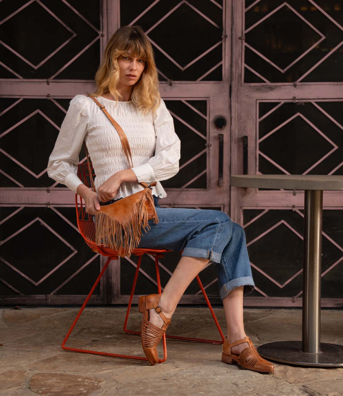 A woman sits outdoors on a red chair, wearing a white blouse, blue jeans, brown woven sandals, and carrying the Bed Stu Silverrock bag. A metal table is nearby with a patterned door in the background.