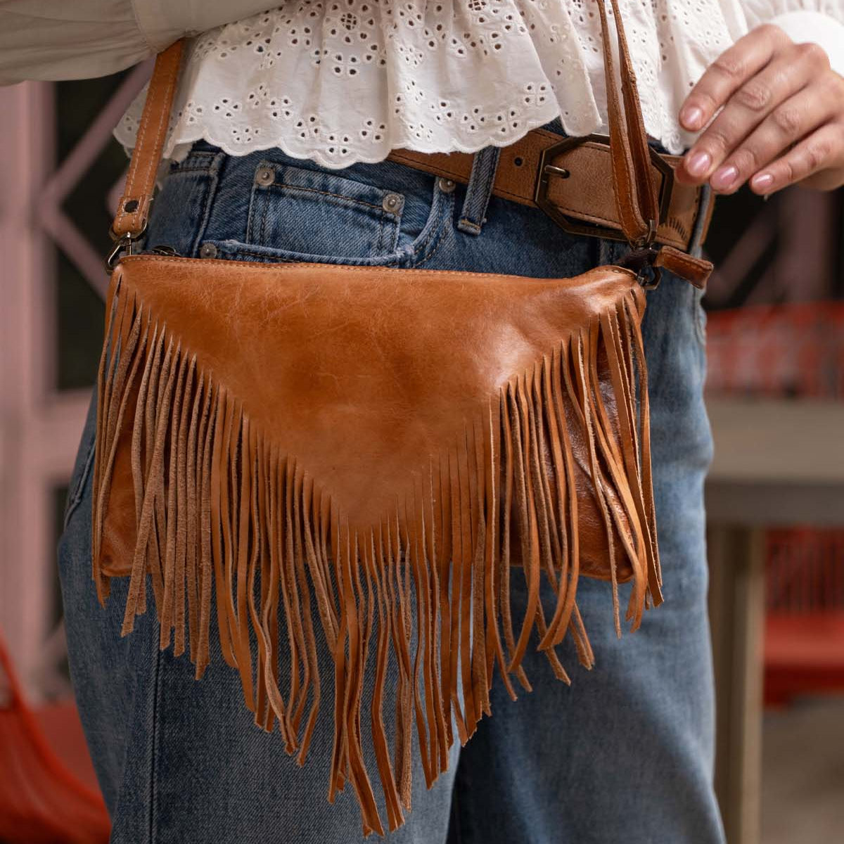 A person wearing blue jeans and a white eyelet blouse carries the Bed Stu Silverrock, a brown leather fringe flap bag with a shoulder strap.