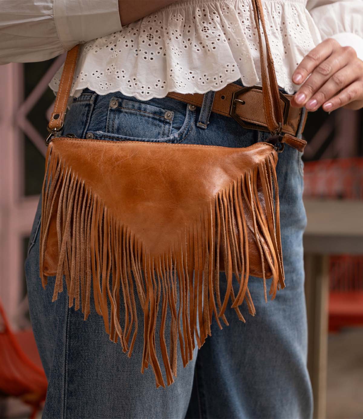 A person wearing blue jeans and a white eyelet blouse carries the Bed Stu Silverrock, a brown leather fringe flap bag with a shoulder strap.