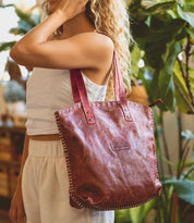 Wearing a white sleeveless top and pants, a person with curly blonde hair carries the Bed Stu Skye II tote—with red handles and visible stitching—making it a stylish, roomy choice for commuting.