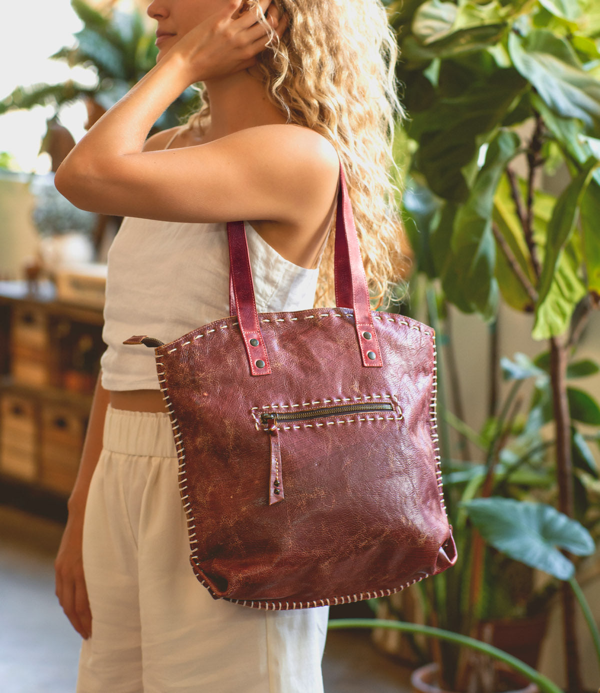 A woman indoors carries the Bed Stu Skye II, a spacious brown leather tote with white stitching and an exterior zip pocket, on her shoulder while wearing a sleeveless white outfit.