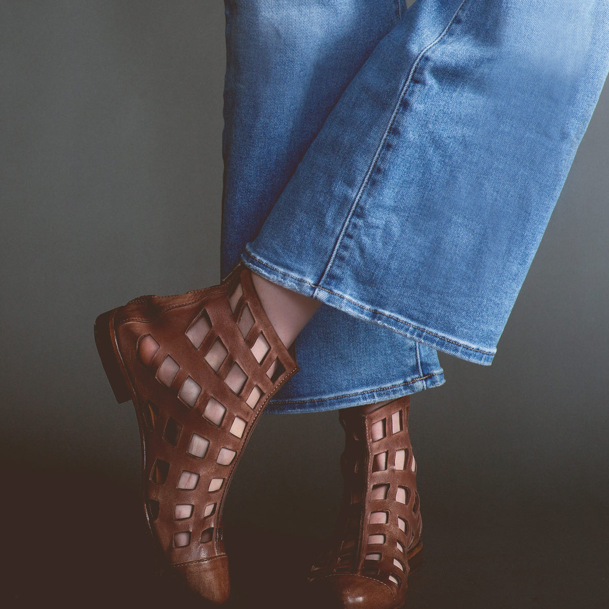 Person wearing flare jeans and **Skyline** brown ankle boots handcrafted in Italy by **Bed Stu**, featuring bold square cutouts, standing with one leg slightly crossed over the other.