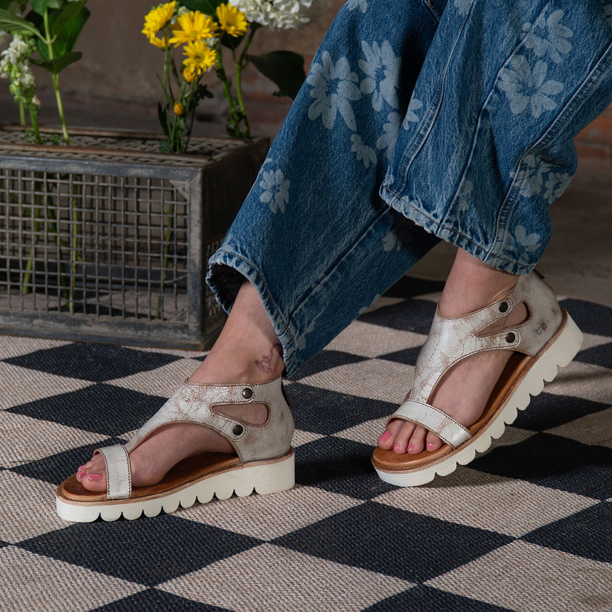 A person in floral jeans and beige Soni II platform sandals by Bed Stu, featuring an XL EXTRALIGHT® outsole, stands on a checkered rug with flowers in a metal box behind them.