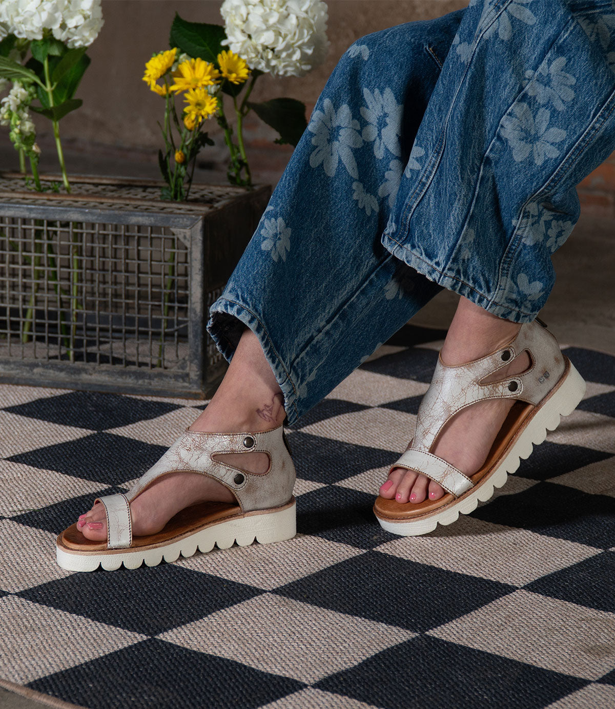 A person in floral jeans and beige Soni II platform sandals by Bed Stu, featuring an XL EXTRALIGHT® outsole, stands on a checkered rug with flowers in a metal box behind them.