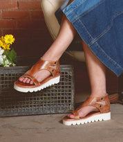 A person wearing Bed Stu's stylish Soni II platform sandals with white XL EXTRALIGHT® outsoles sits with legs crossed next to flowers and a brick wall.