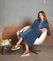 A woman sits in a white chair wearing a denim dress and brown Bed Stu Soni II platform sandals with an XL EXTRALIGHT® outsole. Beside her, a wire crate holds white and yellow flowers against a brick and concrete wall backdrop.