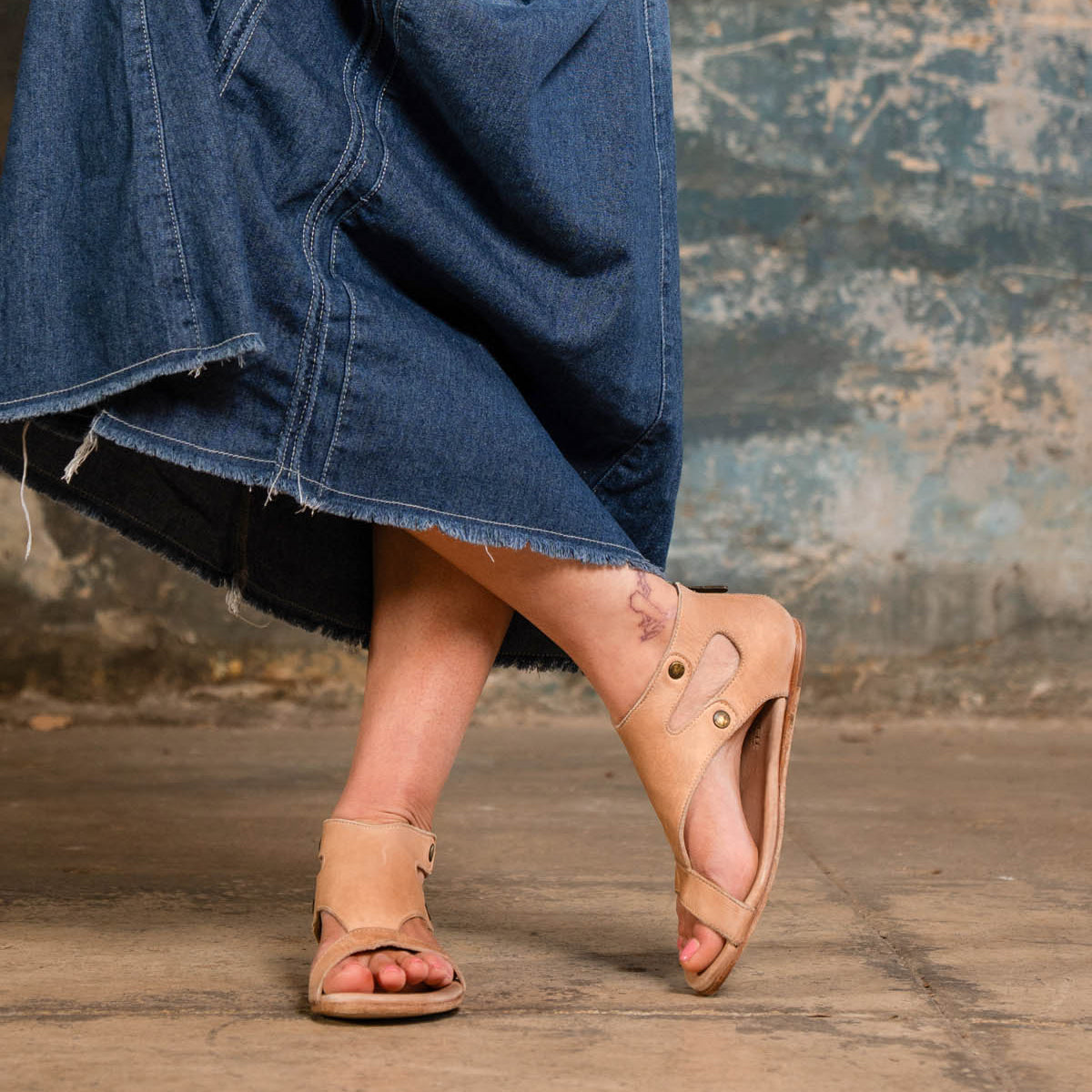 A person wearing a long denim skirt and Bed Stu Soto tan leather sandals with an intricate cutout design stands on a concrete floor against a weathered wall.