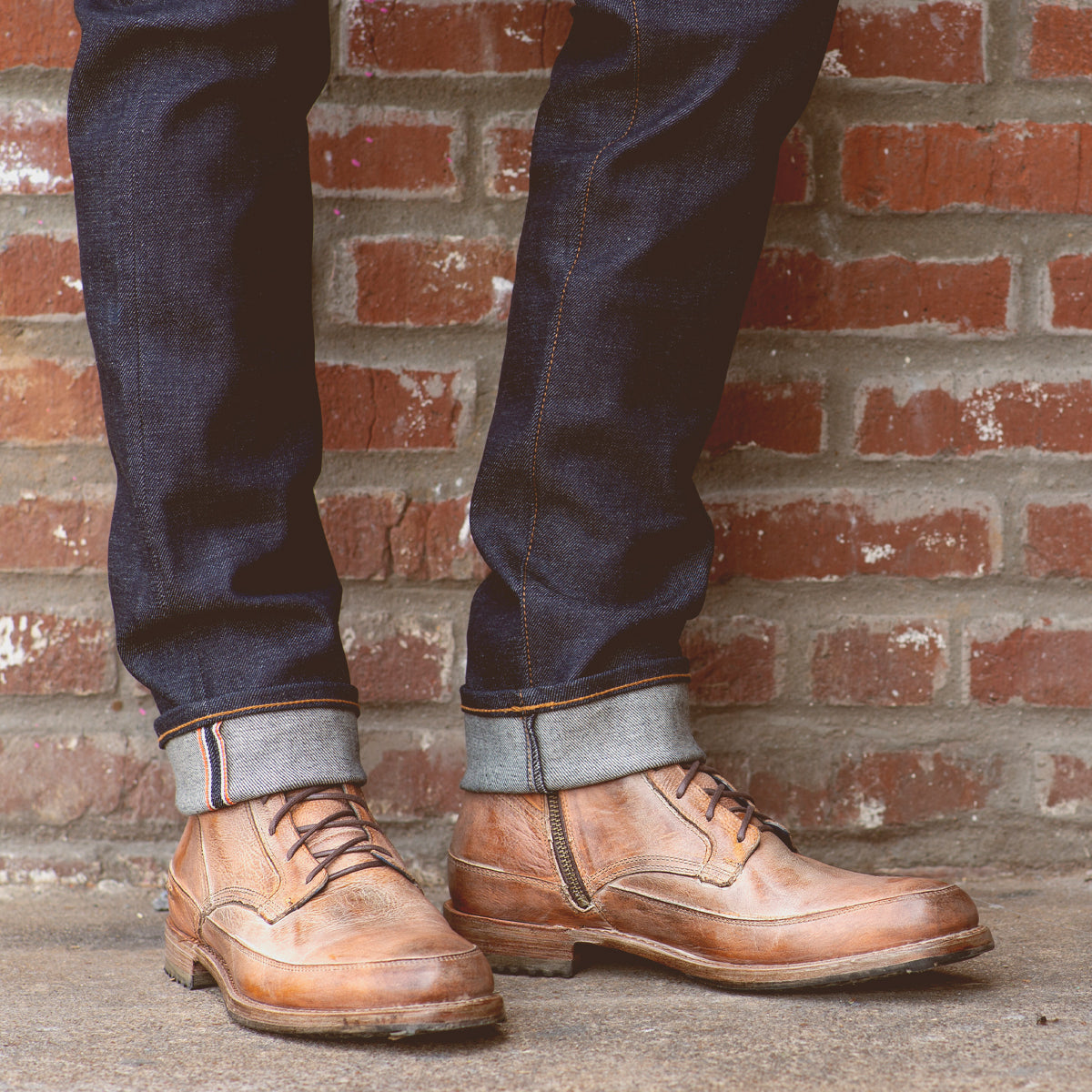 A person wearing Bed Stu Spiker Teak Rustic Boots and cuffed jeans standing against a brick wall.